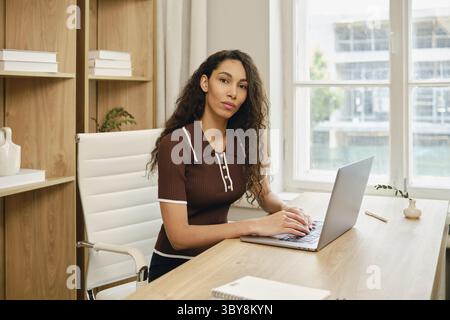 Una donna è seduta su un'elegante scrivania in legno, utilizzando un notebook in un ufficio illuminato dal sole. L'arredamento moderno include luce naturale che scorre attraverso grandi finestre, Foto Stock