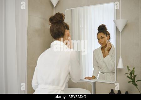 Una donna con l'abito bianco si trova in un bagno elegante e si lava dolcemente il trucco dal viso. Si concentra sul suo riflesso nello specchio, circondato da eleganza Foto Stock