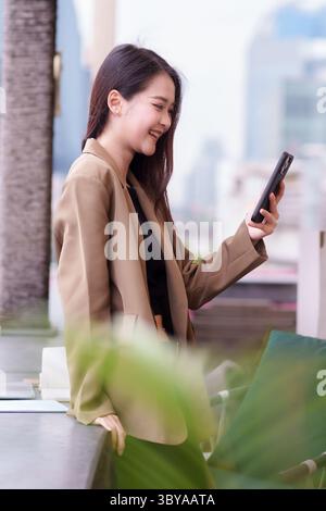 La giovane donna asiatica sorride con fiducia usando il suo dispositivo mobile che si connette digitalmente in un ambiente urbano all'aperto. Abbraccia la tecnologia moderna per le comunicazioni Foto Stock