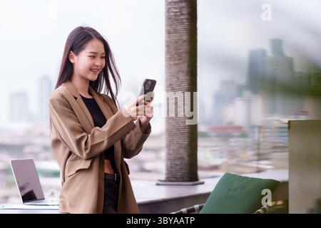 Donna asiatica adulta si erge con sicurezza sul balcone urbano. Si impegna con il suo telefono cellulare. Sfondo urbano moderno con laptop. A una donna professionale piace Foto Stock