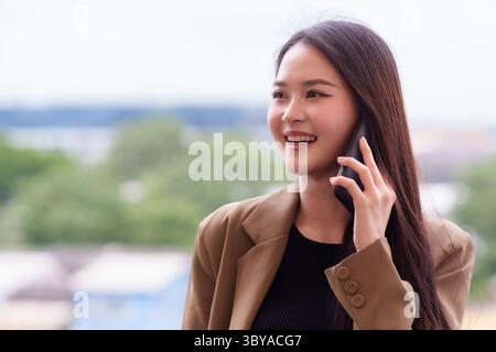Una donna asiatica sorridente si impegna in conversazioni telefoniche all'aperto, mostrando fiducia e professionalità moderna. Questa immagine incarna una virgola efficace Foto Stock