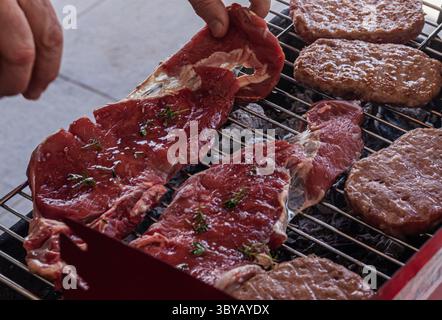 Succose bistecche crude condite con erbe aromatiche e hamburger frizzanti su una griglia a carbone, catturate durante un barbecue sul cortile. Perfetto per un'esperienza visiva culinaria Foto Stock