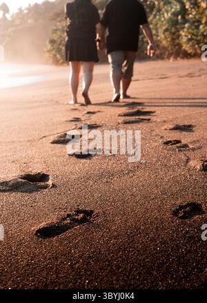 Una coppia romantica cammina a piedi nudi su una spiaggia tropicale al tramonto, lasciando impronte dietro nella sabbia. Foto Stock