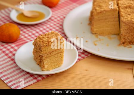 Una fetta di torta al miele a strati con copertura di briciole è elegantemente presentata su un panno a scacchi rosso, accompagnato da arance mandarine, creando un ambiente accogliente e accogliente Foto Stock
