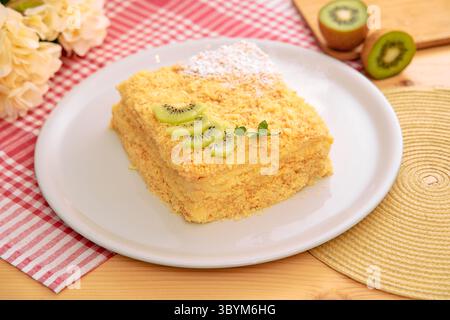 Una fetta di deliziosa torta condita con kiwi fresco e spolverata con zucchero a velo, presentata su un piatto bianco adornato con un tovagliolo a scacchi e un motivo floreale Foto Stock