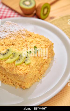 Una deliziosa torta a strati di noci condita con kiwi a fette e un tocco di contorno verde è splendidamente presentata su un piatto bianco, offrendo un delizioso cu Foto Stock