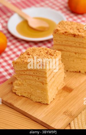 Una deliziosa torta al miele a più strati, ricoperta da una rifinitura friabile, si trova su un asse di legno. è abbinato a un cucchiaio di legno e un piatto di hon Foto Stock