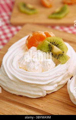 Questa immagine presenta una deliziosa pavlova ricoperta da fresche fette di kiwi e mandarino, adagiata su un asse di legno. perfetto per sfoggiare eleganza nel dessert Foto Stock