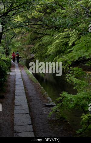 Due turisti che camminano lungo un canale in un tranquillo parco, godendosi la vegetazione lussureggiante e l'atmosfera tranquilla Foto Stock