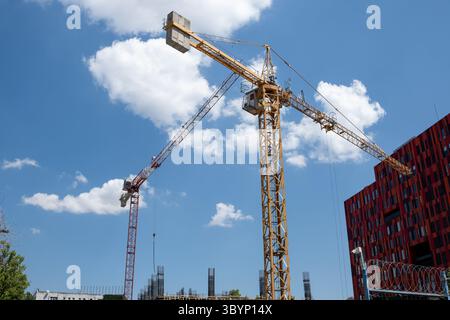 Diverse gru da costruzione stanno lavorando attivamente in un cantiere in un ambiente urbano, con un cielo azzurro limpido e nuvole soffici sopra, che indicano a poppa Foto Stock