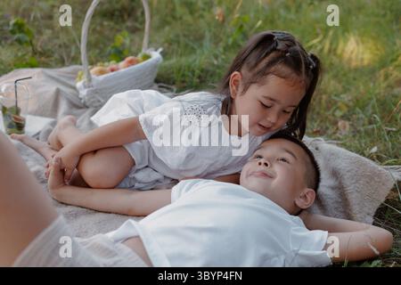 Due bambini si rilassano su una coperta da picnic nell'erba, condividendo un momento dolce durante una giornata estiva con la famiglia Foto Stock
