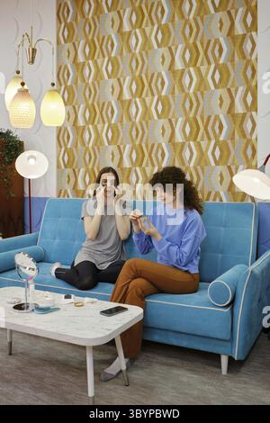 Due amici si stanno godendo un momento di relax su un divano blu, impegnati in una sessione di bellezza. Stanno applicando cerotti e panna mentre ridono e condividono Foto Stock