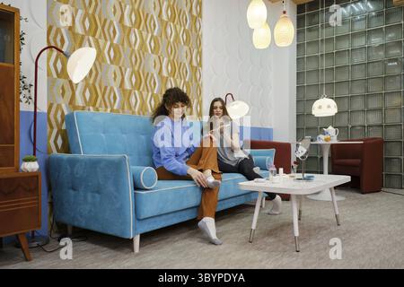 Due donne sono sedute su un divano blu in un soggiorno, godendosi la compagnia dell'altra mentre si impegnano in procedure di bellezza Foto Stock