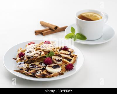 Waffle belgi con lamponi, banana, sciroppo di cioccolato. Colazione con tè su sfondo bianco, vista laterale Foto Stock