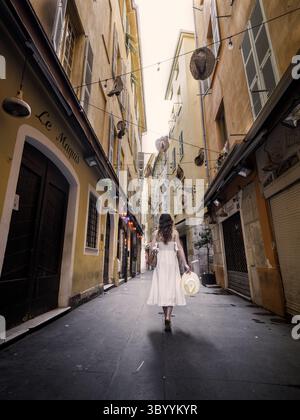 Donna in abito bianco che cammina attraverso la stretta strada vecchia decorata con cappelli appesi nel centro storico di Nizza, Provenza, Francia. Foto Stock