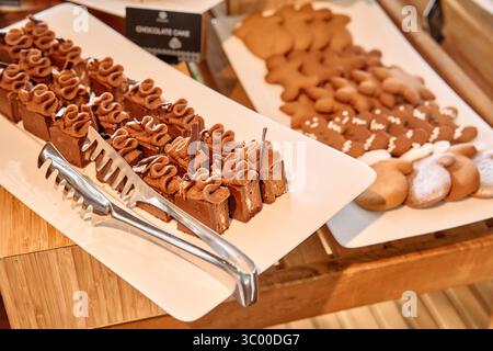Piatti rettangolari di torte al cioccolato e biscotti al pan di zenzero, guarniti con glassa, esposti su un buffet con pinze, che offre un dolce e festoso Foto Stock