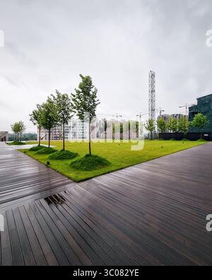 Piccolo parco nel quartiere degli affari. Passeggiata sul lungomare che circonda un prato con erba verde brillante e piccoli alberi. Foto Stock