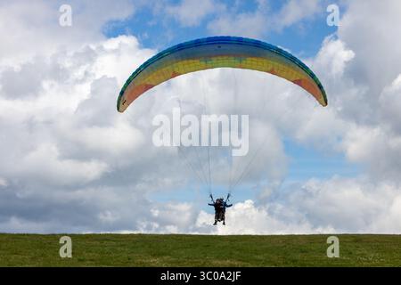 Vista di un parapendio che si innalza in alto su uno sfondo di nuvole bianche e un cielo blu vibrante, sospeso su un lussureggiante paesaggio verde, Oderen, Grand Est, Francia. Foto Stock
