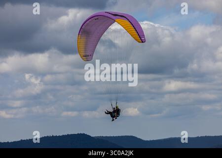 Vista di un vibrante parapendio che vola attraverso il cielo, una danza di viola e giallo sullo sfondo di nuvole fluttuanti, Oderen, Grand Est, Francia. Foto Stock