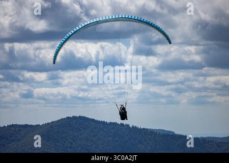 Vista di un parapendio che si innalza sullo sfondo di cieli spettacolari e striati di nuvole e colline ondulate e boscose, Oderen, Grand Est, Francia. Foto Stock