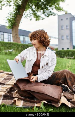 La giovane donna con i capelli ricci lavora in remoto sul suo notebook in un parco soleggiato. Foto Stock