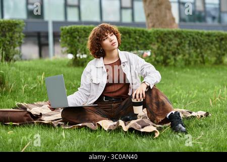 Una donna dai capelli ricci lavora sul suo laptop, circondata da erba verde, che abbraccia le vibrazioni estive all'aperto. Foto Stock