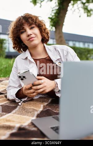 Una donna dai capelli ricci lavora al telefono da una coperta accogliente in un parco verde lussureggiante sotto il sole. Foto Stock