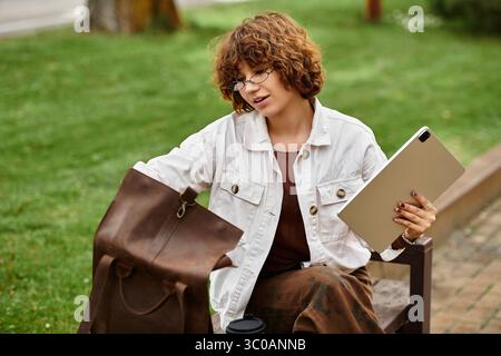 Una donna dai capelli ricci lavora in remoto in un parco soleggiato, bilanciando laptop e caffè. Foto Stock