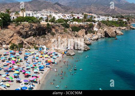 Vista aerea di una spiaggia affollata, Playa Carabeillo, con ombrelloni colorati e nuotatori nelle acque turchesi vicino alle scogliere rocciose. Nerja, Spagna Foto Stock