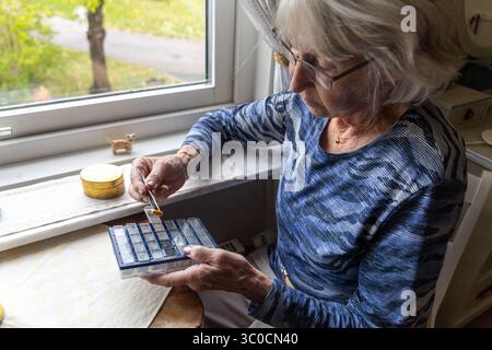 Un'anziana donna dai capelli grigi organizza meticolosamente le sue pillole di medicina con delle pinzette, sedute vicino alla finestra nel comfort della propria casa. Foto Stock