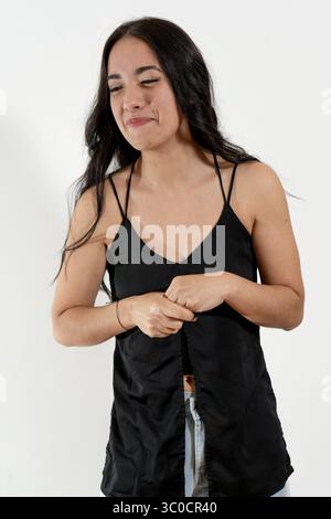 Giovane donna che si imbatte con la sua camicetta, esprime disagio, timidezza o insicurezza su sfondo bianco Foto Stock