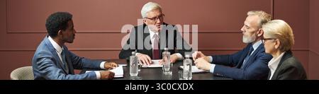 Gruppo di politici di mezza età e anziani tra cui uomo nero, uomo caucasico, donna caucasica seduta a tavola a discutere documenti durante una riunione formale in ufficio Foto Stock