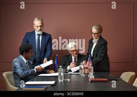 Gruppo di politici di mezza età e anziani, tra cui uomo nero, uomo caucasico, donna caucasica che esamina e firma documenti al tavolo delle conferenze con bandiere europee e americane visibili Foto Stock