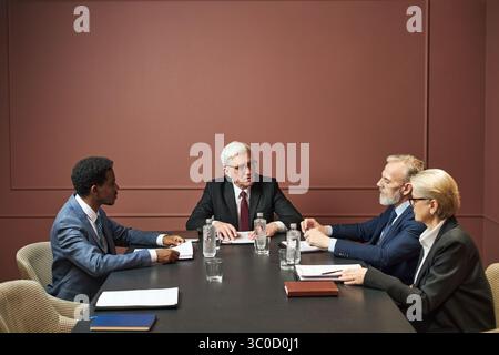 Gruppo di politici di mezza età e anziani, tra cui uomini neri e caucasici e donne seduti al tavolo di conferenza a discutere documenti durante l'incontro formale Foto Stock