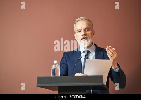 Uomo caucasico di mezza età in piedi sul podio che pronuncia discorsi, tiene la carta in una mano e gesti con l'altra, bottiglia d'acqua sul podio, guardando leggermente oltre la fotocamera Foto Stock
