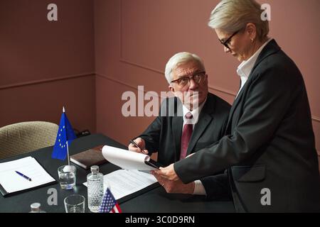 Anziano uomo caucasico seduto a tavola firmando documenti mentre donna caucasica di mezza età in piedi accanto a lui in possesso di documenti, entrambi indossano abiti da lavoro Foto Stock