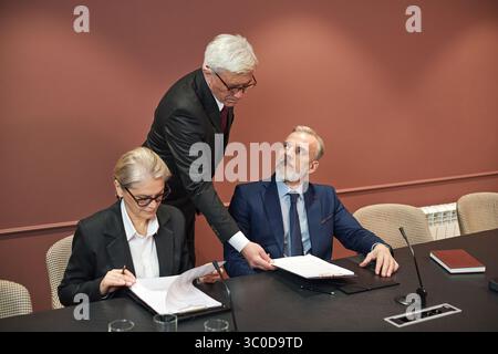 Uomo caucasico di mezza età in piedi, consegnando un documento a una donna caucasica di mezza età seduta e a un uomo caucasico di mezza età durante una riunione di lavoro nella sala conferenze Foto Stock
