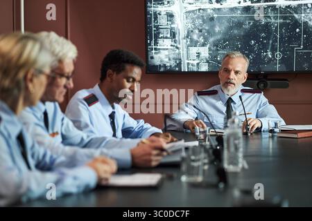 Gruppo di politici di mezza età e anziani, tra cui uomini e donne caucasici e neri seduti al tavolo delle conferenze, che discutono di strategia con una grande mappa visualizzata sullo schermo Foto Stock