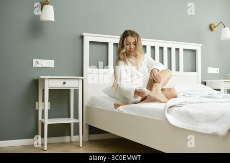 Una donna di mezza età siede su un letto in una camera da letto moderna che si stacca e rimuove la pelle morta dai suoi piedi Foto Stock