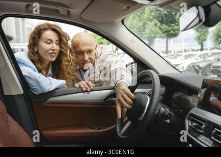 Una coppia esamina gli interni di un veicolo guardando attraverso il finestrino aperto di una concessionaria, discutendo le caratteristiche e le opzioni Foto Stock