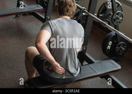 Atleta o giovane uomo seduto con dolore nella parte bassa della schiena in palestra con tensione muscolare. Braccio, infiammazione o esercizio fisico con disagio nel centro fitness. Clos Foto Stock