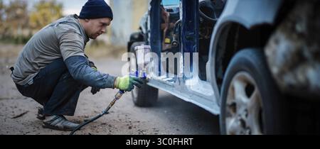 Un uomo che accovacciava, dipingeva un'auto con una pistola a spruzzo all'aperto Foto Stock
