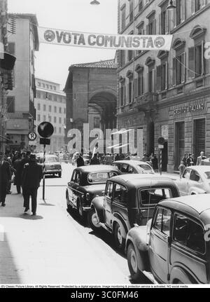 Italia negli anni '1950 Una strada nella città di Firenze con la Loggia del mercato nuovo sullo sfondo. Le auto Fiat 600 risalenti all'anno 1955 sono parcheggiate lungo la strada. Di fronte alla strada è appeso uno striscione con il testo "Vota Comunista” (Vota Comunista). Le elezioni nazionali in Italia negli anni '1950 si sono svolte nel 1953 e nel 1958. Tuttavia, le elezioni locali o le campagne politiche avrebbero potuto avvenire nel 1955, e la bandiera "Vota Comunista" suggerisce un'attività politica comunista attiva, che fu forte in Italia durante questo periodo, soprattutto in città come Firenze. La Fiat 600, introdotta nel 195 Foto Stock