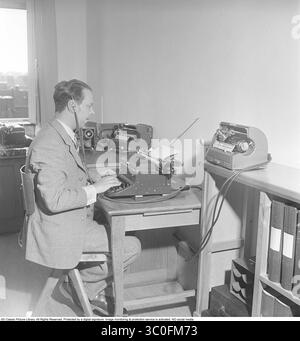 La fotografia in bianco e nero scattata nel 1949 raffigura un uomo seduto su una scrivania di legno, che scrive su una macchina da scrivere ciò che sente mentre ascolta la registrazione su un apparecchio Ediphone con le cuffie. L'Ediphone era una prima macchina di dettatura sviluppata dalla compagnia di Thomas Edison, progettata per registrare e riprodurre parole pronunciate a scopo di trascrizione. Funzionava utilizzando un cilindro di cera come mezzo di registrazione, sul quale il suono era inciso da uno stilo collegato a un diaframma che vibrava con la voce dell'altoparlante. L'utente parlava in un boccaglio e la macchina acquisiva l'audio. A. Foto Stock