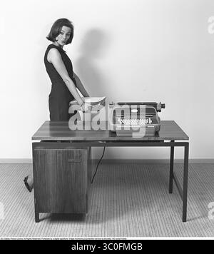 Il Friden 5010 Computyper 1965. L'immagine raffigura una giovane donna in piedi accanto a una scrivania di legno, che tiene una pila di carte. Sulla scrivania si trova un Friden 5010 Computyper, una macchina per la digitazione elettronica e il calcolo lanciata dall'azienda americana Friden. Non era un computer, ma era dotato di una funzione di calcolatrice e di una tastiera collegata elettricamente a una stampante. Il Friden 5010 Computyper, della società americana Friden, era un'innovativa macchina per la digitazione elettronica e il calcolo progettata per l'uso aziendale. Non era un vero computer, ma combinava una funzione di calcolatrice con una tastiera lin elettrica Foto Stock