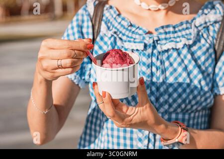 Il turista si gusta un delizioso gelato ai frutti di bosco in una tazza di carta con un cucchiaio piccolo, in una soleggiata giornata estiva Foto Stock