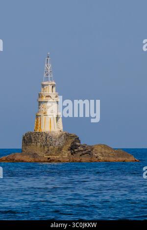 Un faro storico si erge alto su un affioramento roccioso in mare aperto, circondato da tranquille acque blu sotto un cielo limpido, a illustrare la bellezza di Foto Stock