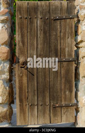 Una robusta porta di legno, intemprata e rustica, è adagiata all'interno di una parete di pietra splendidamente realizzata. La porta è dotata di ferro visibile e si distingue in un tra Foto Stock