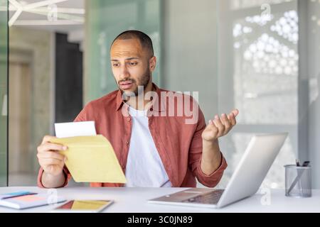 Un uomo sembra confuso mentre guarda una lettera, seduto a una scrivania con un portatile di fronte. Foto Stock