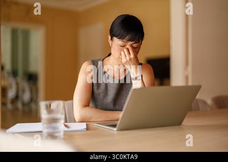 Una donna d'affari stanca che tocca la testa e sta subendo un esaurimento mentre lavora con un notebook alla scrivania aziendale Foto Stock
